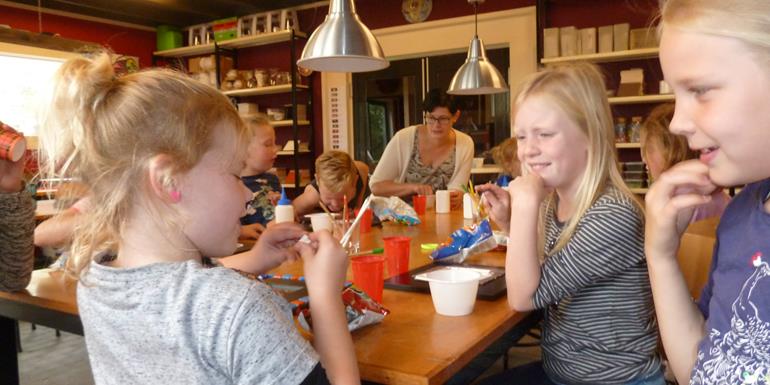 Kinderfeestje Drenthe drie kinderen zitten met elkaar te overleggen, hoe zal ik dit steenje eens opplakken. Op de achtergrond zie je stellingen met mozaiek tegels
															en de grote werktafel met twee lampen